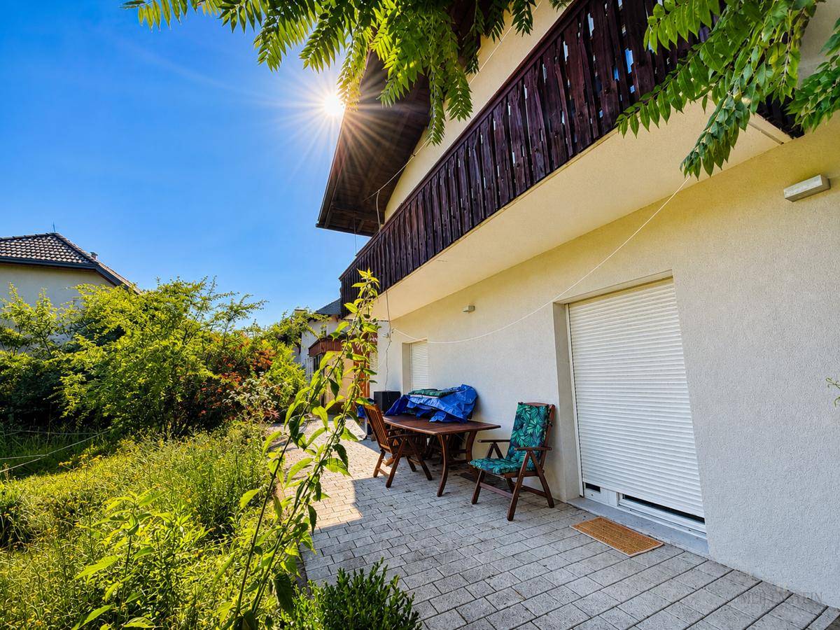 Sonniger Terrassenbereich mit Sitzgelegenheiten und einem Balkon darüber, umgeben von viel Grün.
