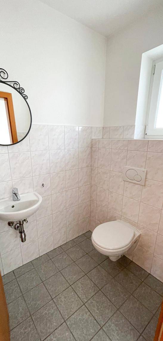 Gäste-WC mit wandhängender Toilette, kleinem Waschbecken und Fenster für Tageslicht.
