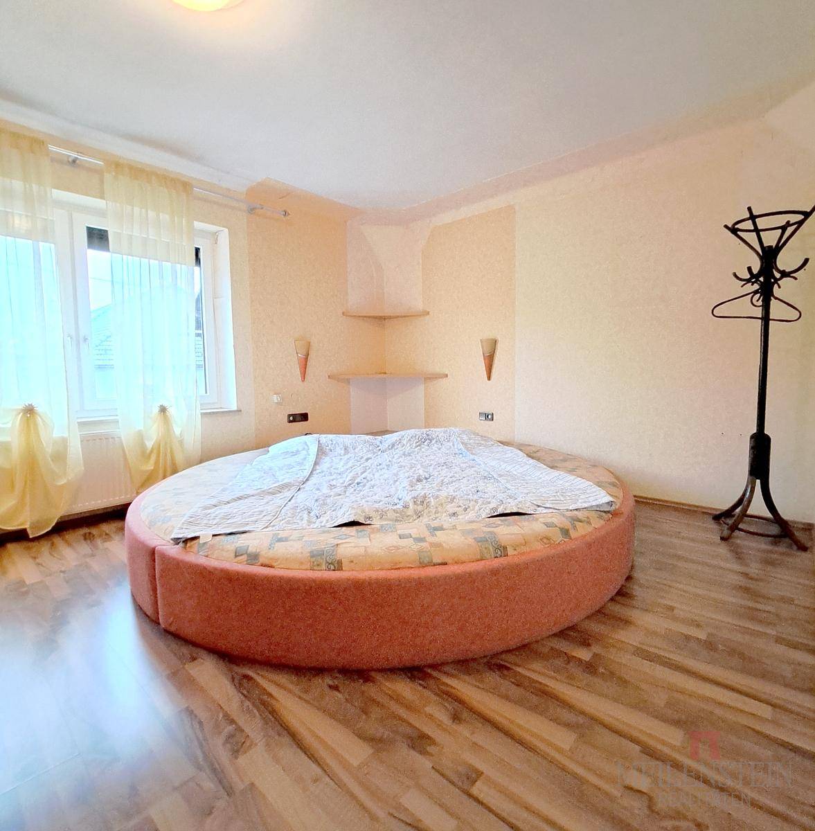Schlafzimmer mit rundem Bett, Laminatboden und hellen Wänden, gut beleuchtet.