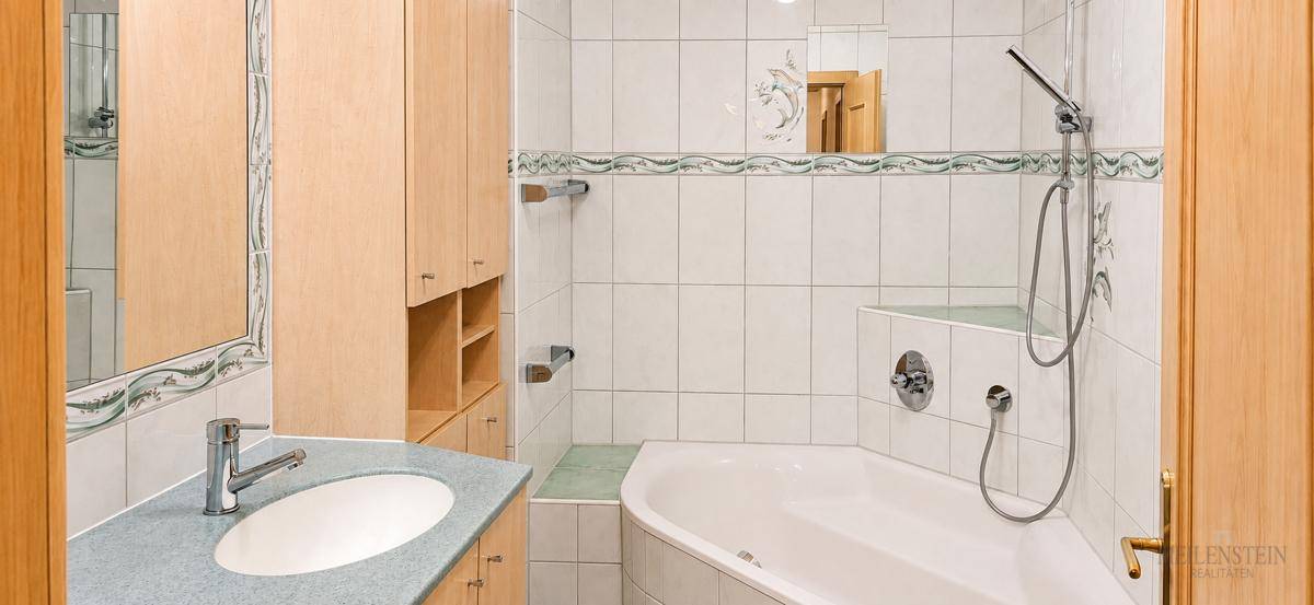 Badezimmer mit Eckbadewanne, Einzelwaschbecken, Spiegelschrank und hellen Fliesen mit Zierleiste.