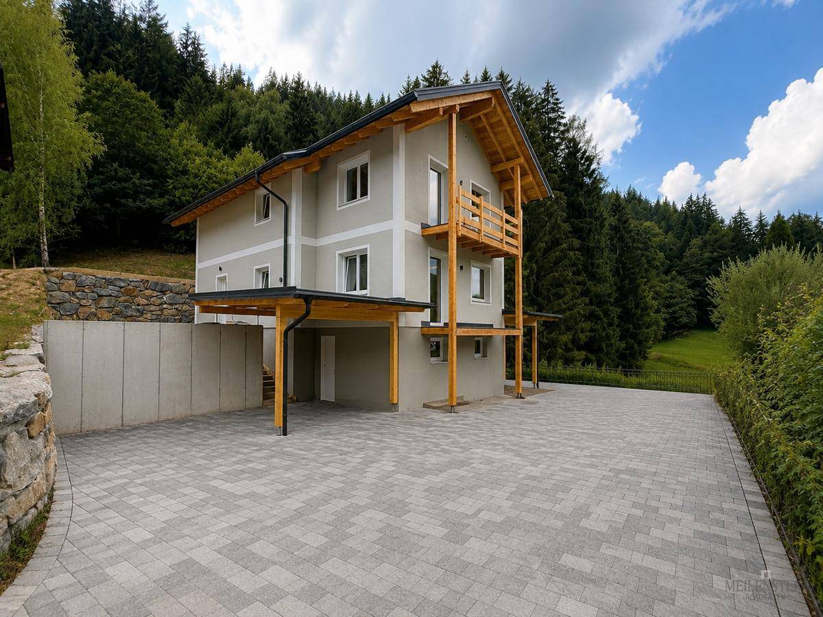 Neubau-Einfamilienhaus in Hanglage mit Holzbalkonen, Carport und gepflasterter Zufahrt, umgeben von Wald.