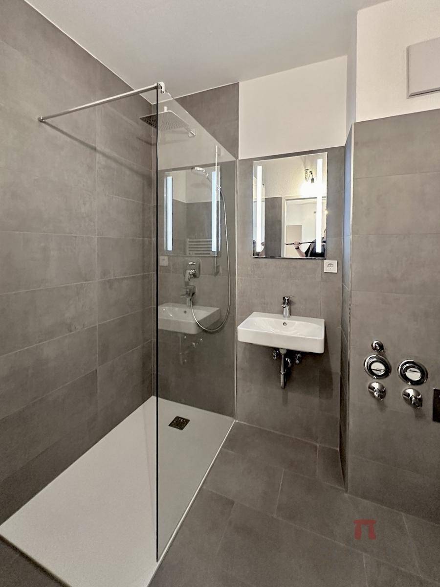 Modernes Badezimmer mit großformatigen grauen Fliesen, begehbarer Dusche und Waschbecken.