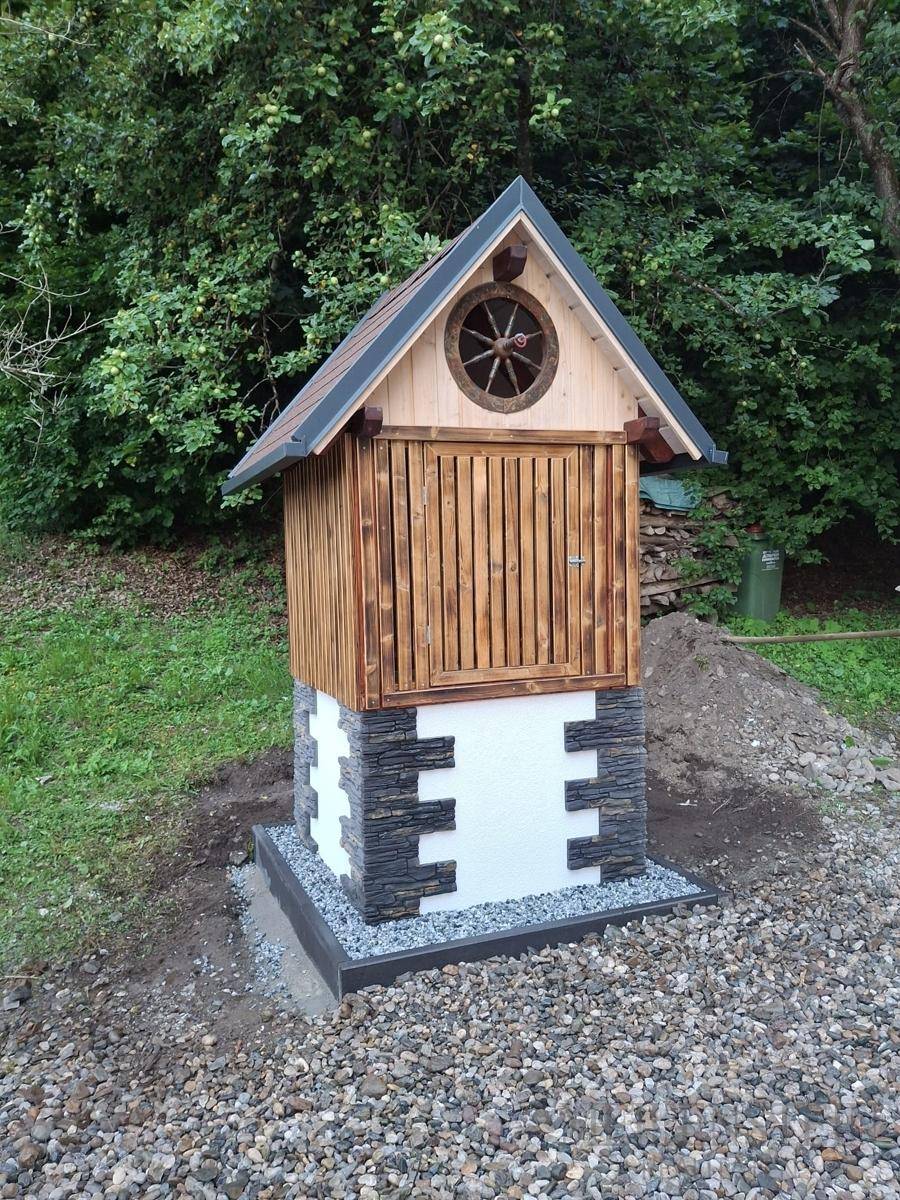 Kleines dekoratives Holzhaus mit Steinsockel und Spitzdach, verziert mit einem Wagenrad im Giebel, umgeben von Kies.