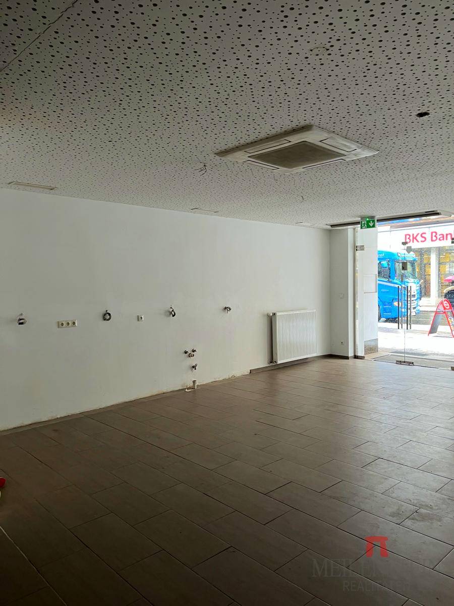 Geräumiger Gewerberaum mit Fliesenboden, Heizkörper und Anschlüssen an der Wand, bereit für den Ausbau.
