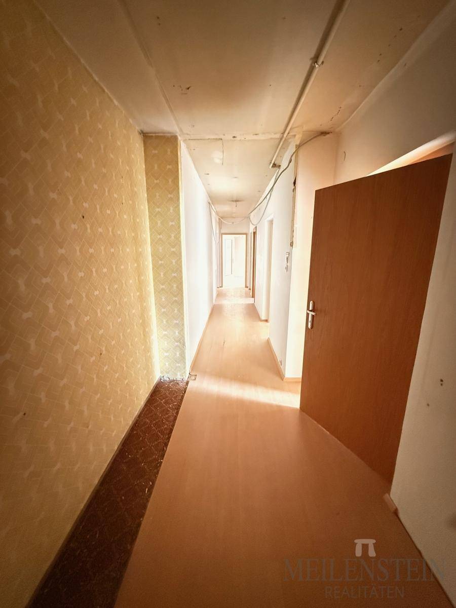Langer Flur mit Linoleumboden und teilweise abgelöster Tapete, der Renovierungsbedarf aufweist.