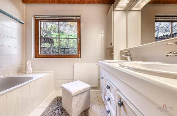 Helles Badezimmer mit Badewanne, Doppelwaschbecken und Fenster mit Blick ins Grüne.
