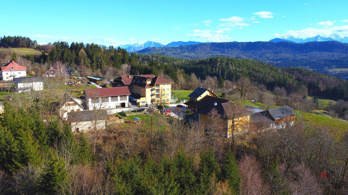 Panoramablick auf eine ländliche Siedlung mit Häusern und bewaldeten Hügeln im Hintergrund.