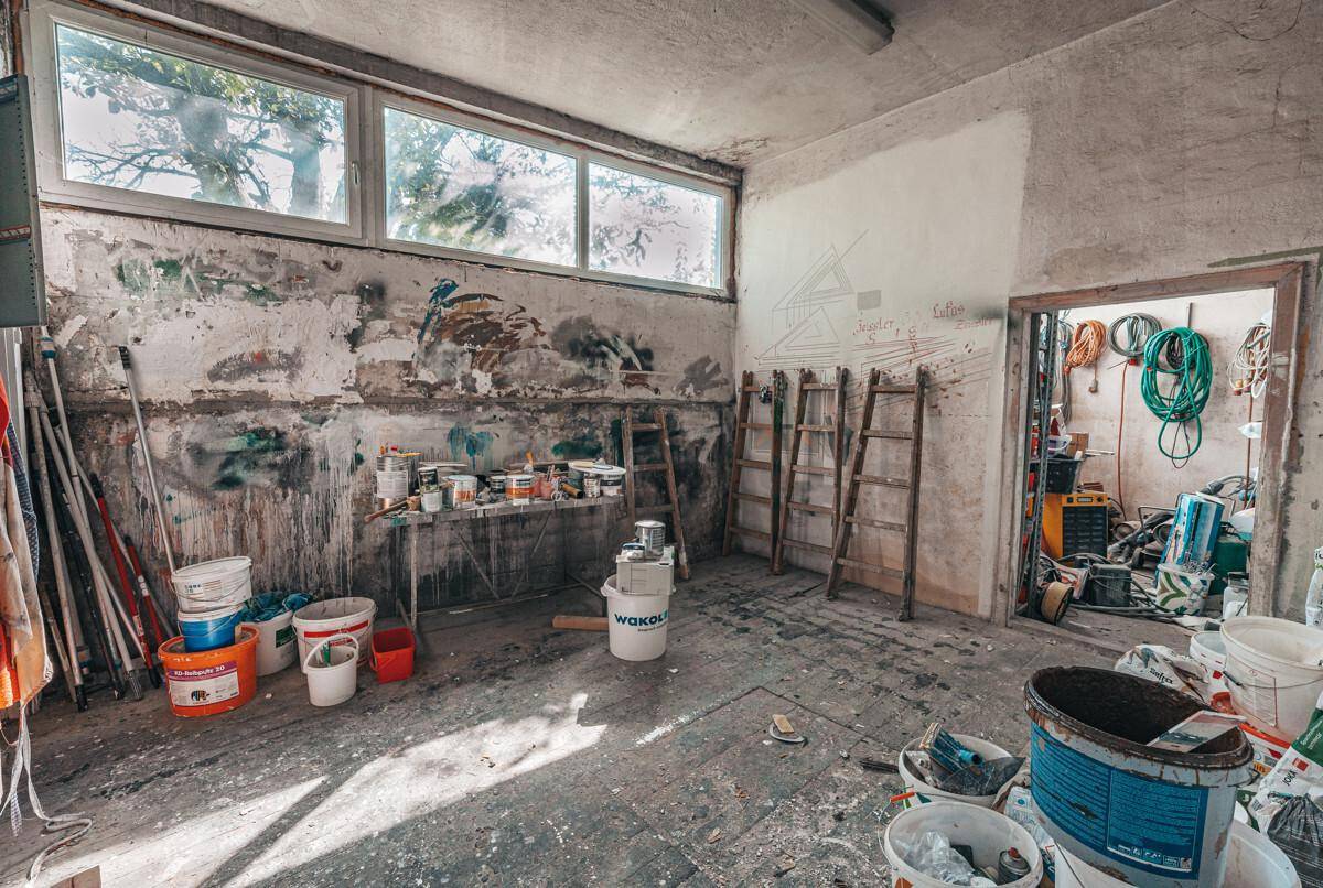 Werkstattraum mit großen Fenstern, Farbeimern und Leitern, bereit für Renovierungsarbeiten.