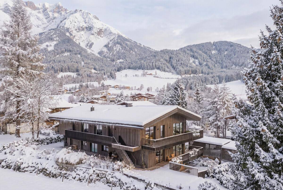 Panoramablick auf das moderne Chalet mit Holzfassade und Balkonen in verschneiter Berglandschaft.