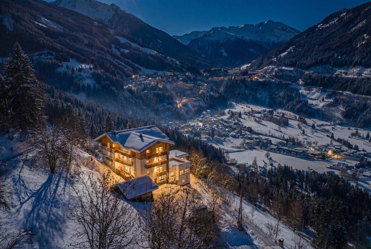Nachtaufnahme eines beleuchteten Gebäudes in einem verschneiten Tal, umgeben von Bergen.