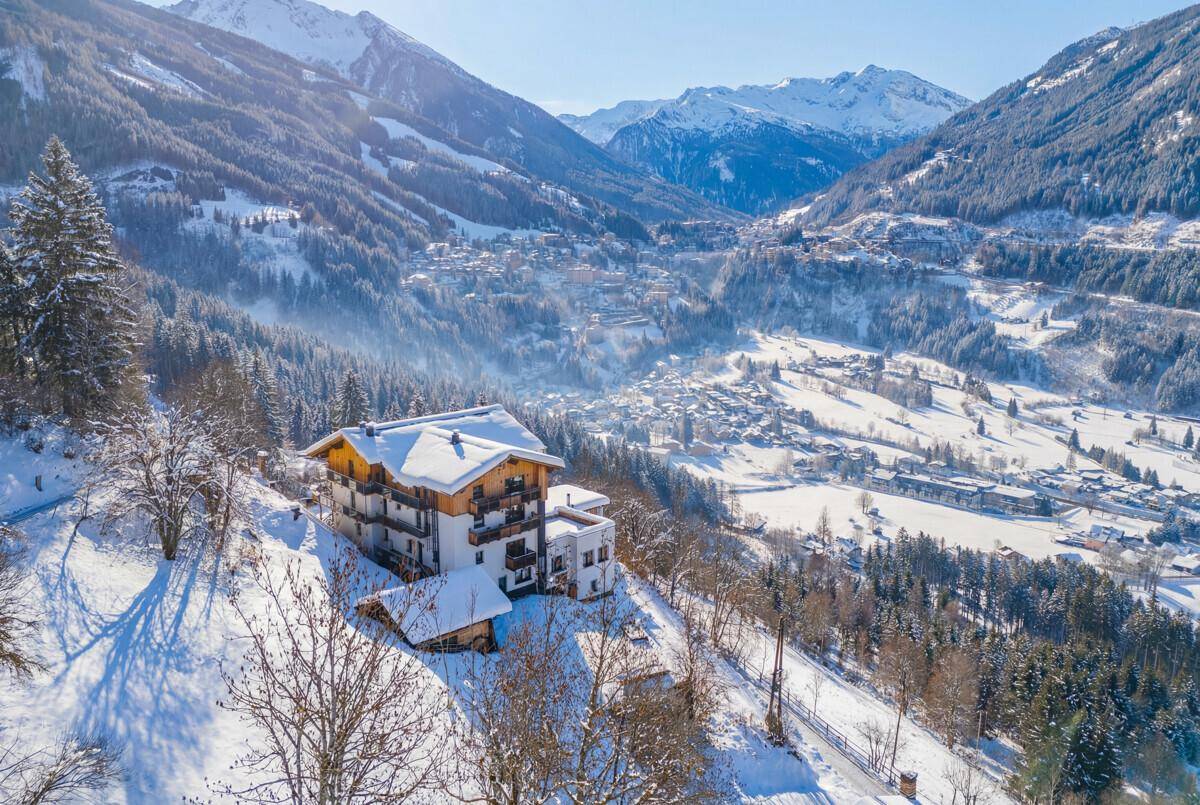 Panoramablick auf ein Gebäude in einem verschneiten Tal, umgeben von Bergen und einem Dorf in der Ferne.