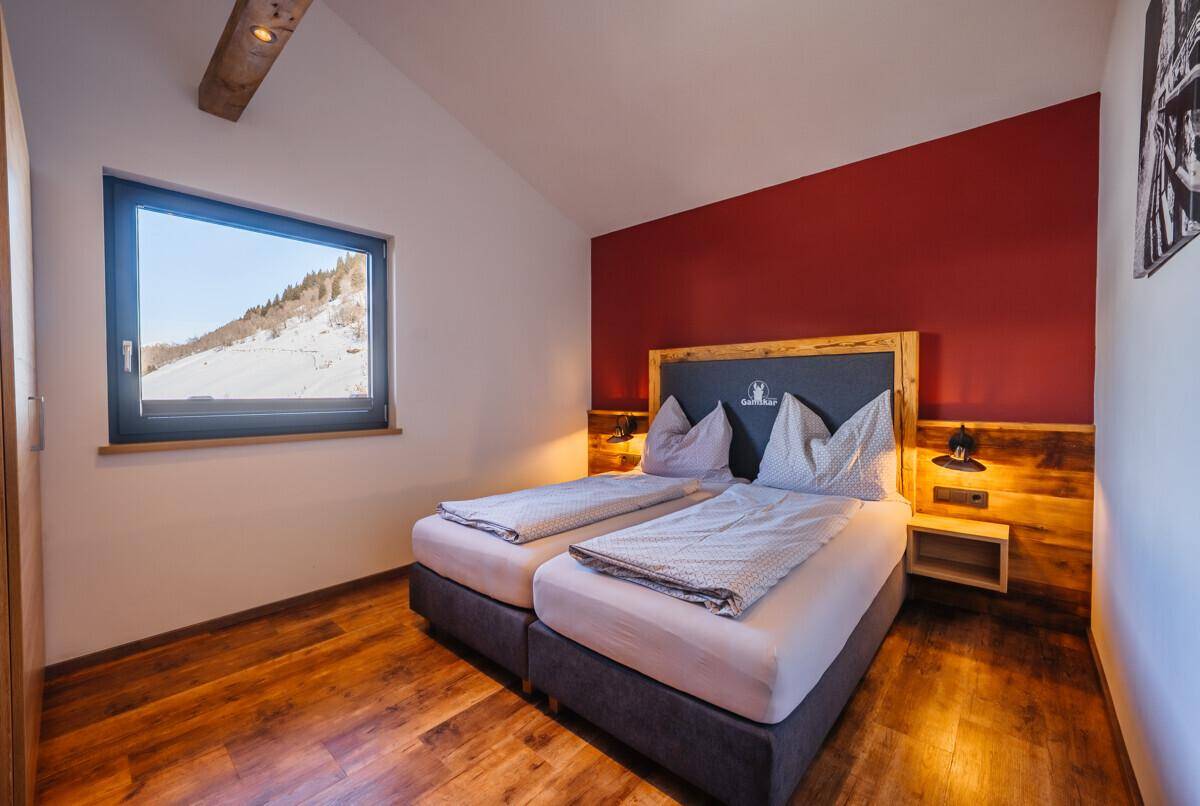 Schlafzimmer mit Doppelbett, roter Akzentwand und Fenster mit Blick auf die verschneiten Berge.