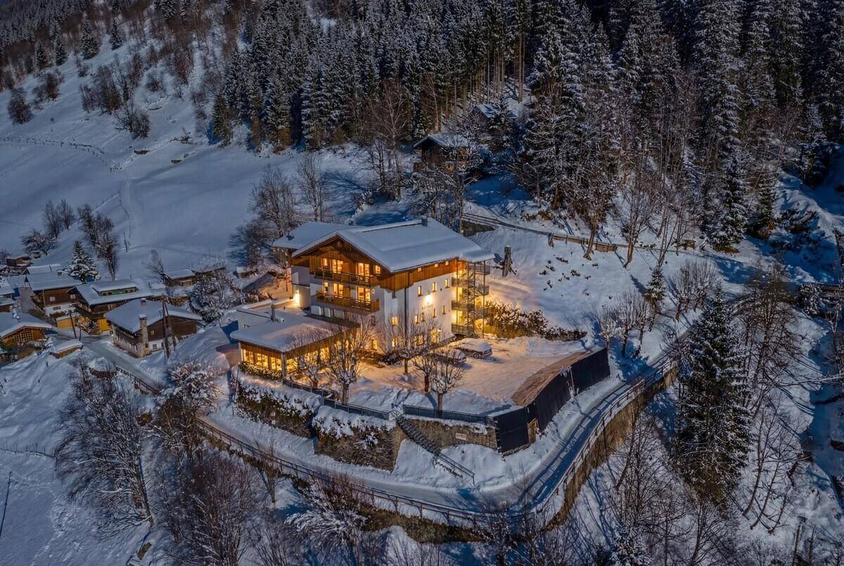 Luftaufnahme eines beleuchteten Gebäudes in verschneiter Berglandschaft bei Dämmerung mit vielen Bäumen.