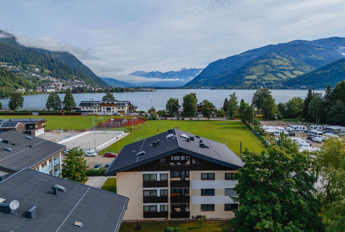 Luftaufnahme des Gebäudes mit Blick auf den See, Berge und Sportanlagen in einer malerischen Landschaft.