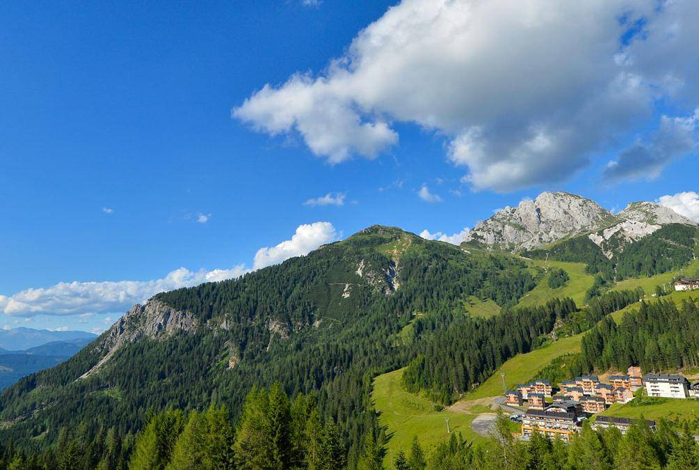 Weitläufige Berglandschaft mit grünen Hängen, dichten Wäldern und einer Ferienanlage unter blauem Himmel.