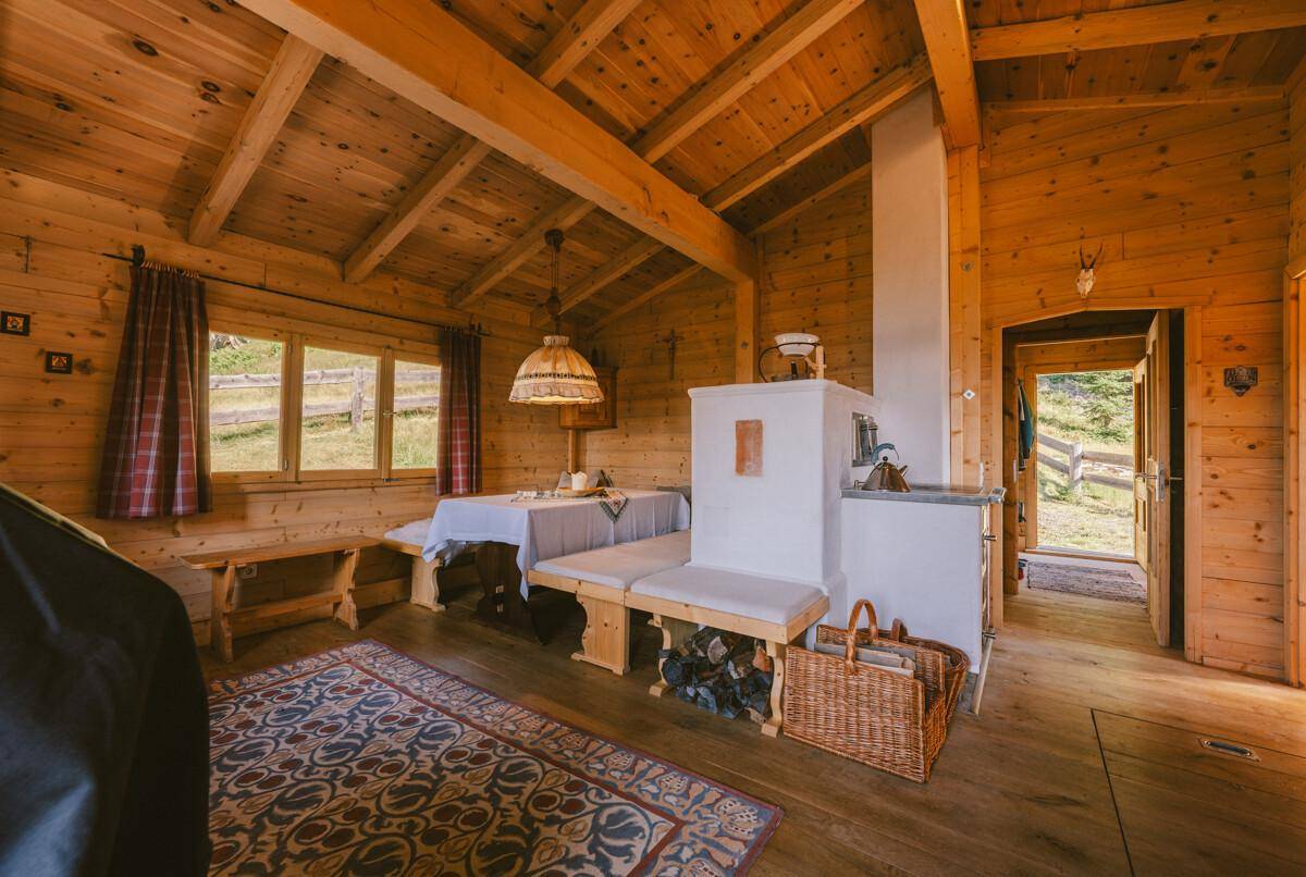 Gemütlicher Wohn-Essbereich in einer Holzhütte mit traditionellem Kachelofen und Holzbänken.