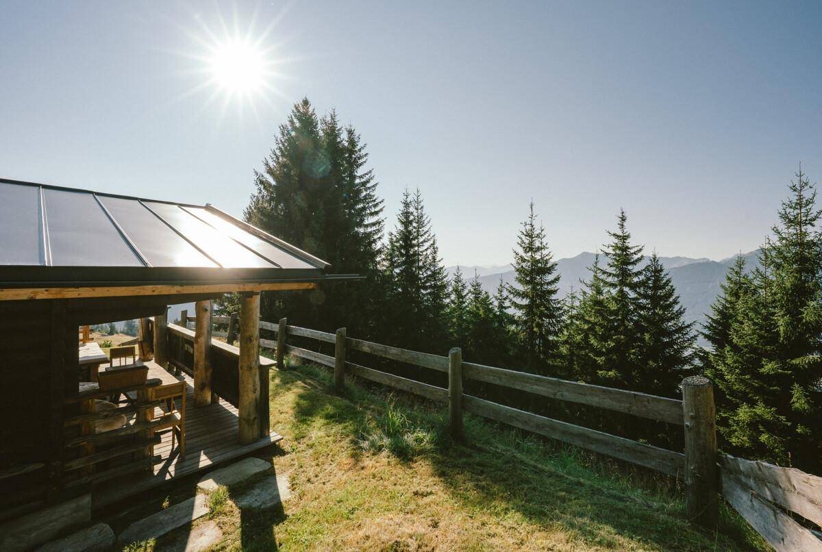 Sonnige Terrasse mit Holztisch und Bänken, umgeben von alpiner Natur und mit Solaranlage.