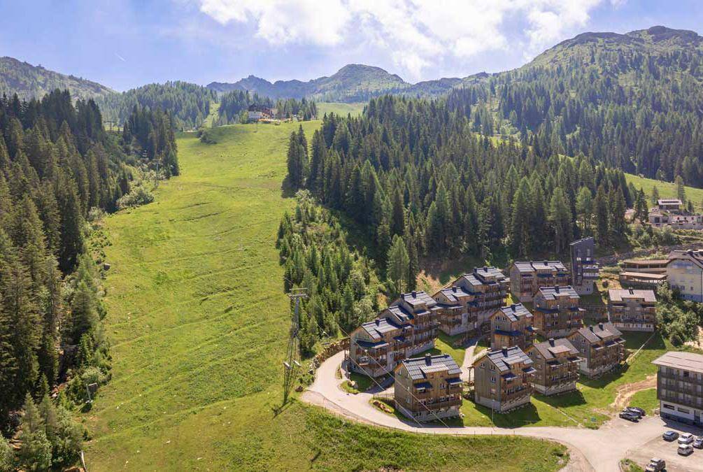 Luftaufnahme einer Ansammlung von Gebäuden in einer grünen, bewaldeten Berglandschaft im Sommer.