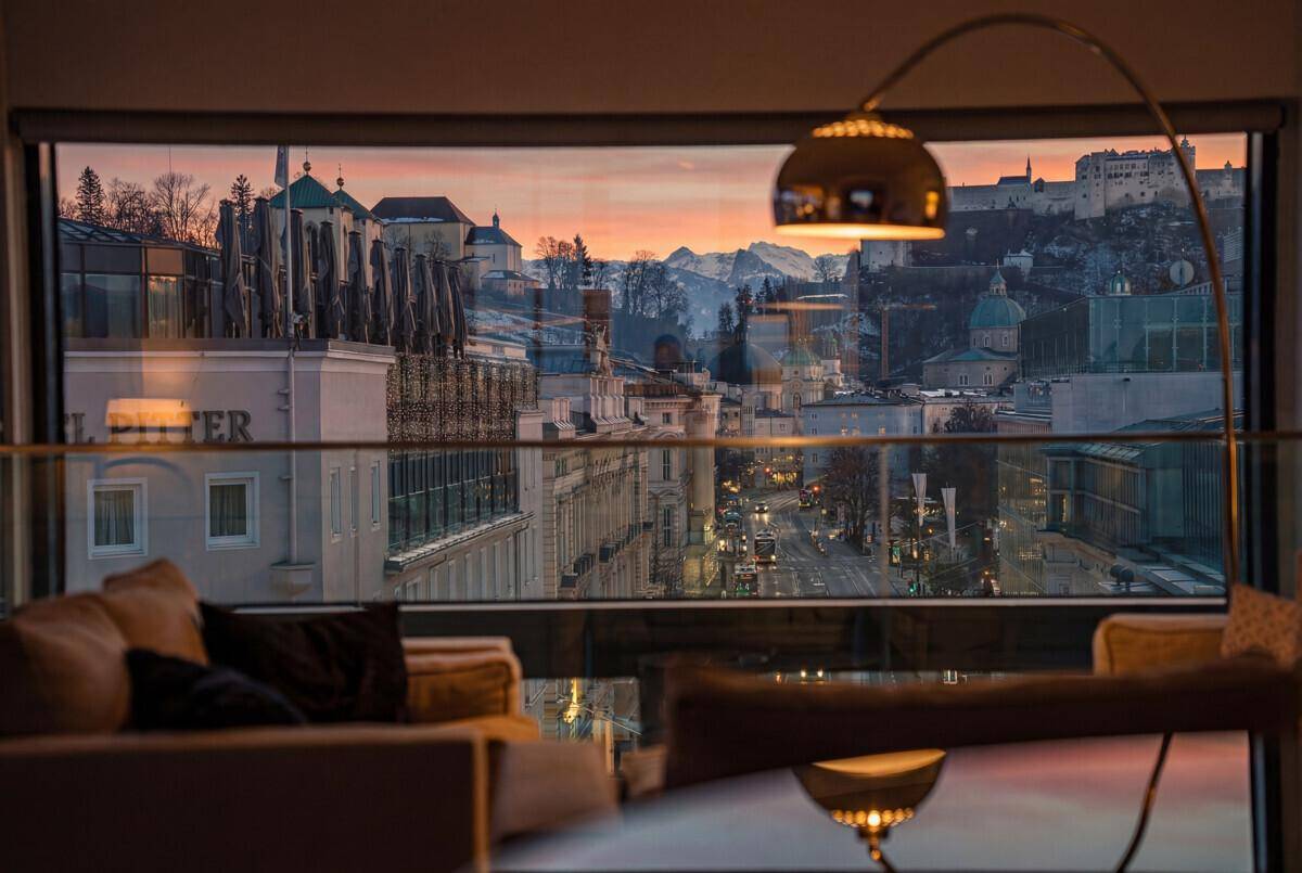 Gemütliches Wohnzimmer mit großem Fenster und Panoramablick auf die Stadt und Berge bei Sonnenuntergang.