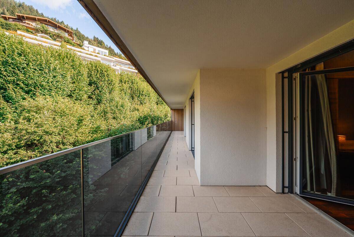 Langer Balkon mit Fliesenboden und Glasgeländer, umgeben von üppigem Grün und Bergblick.