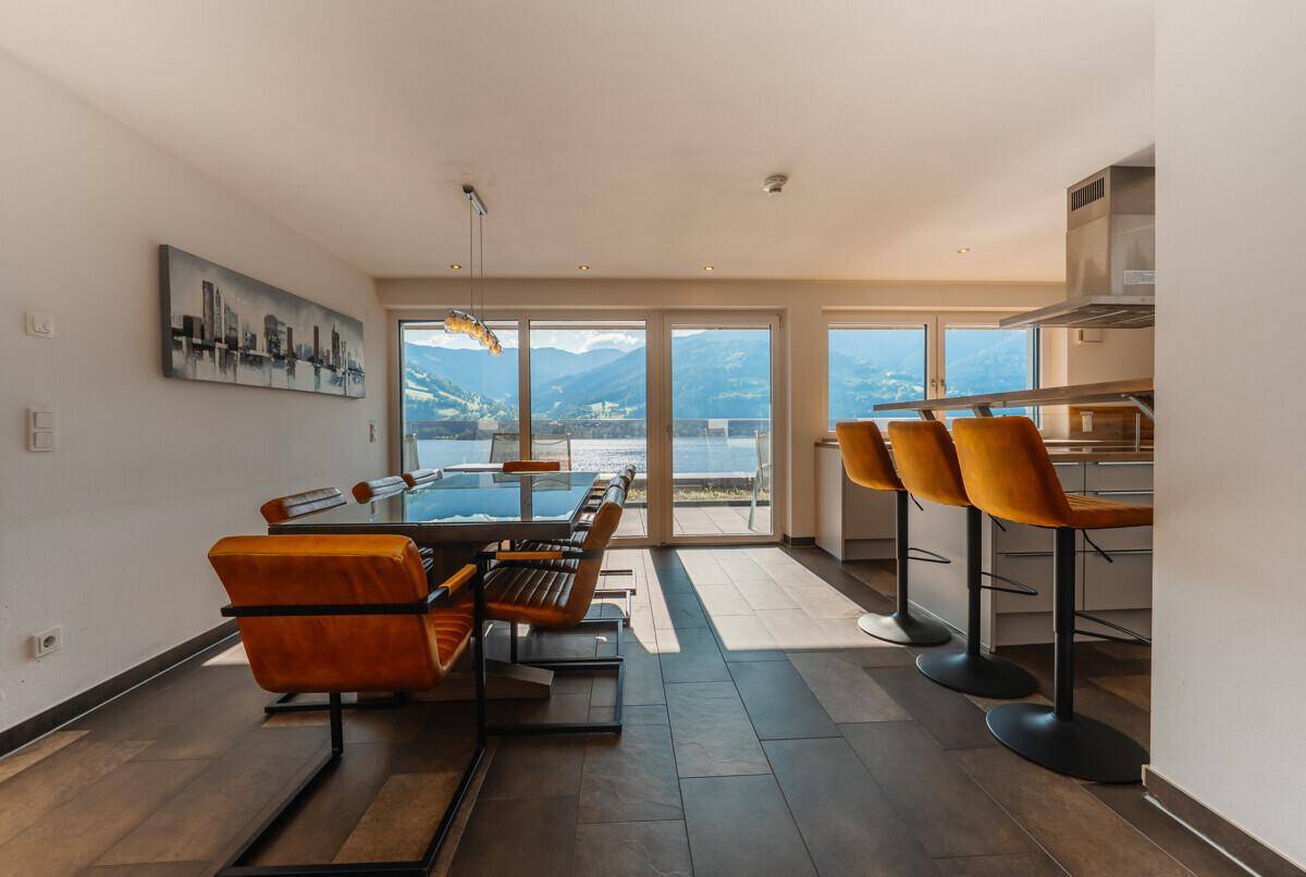 Moderner Essbereich mit orangefarbenen Stühlen und Barhockern, angrenzend an die Küche mit Bergblick.
