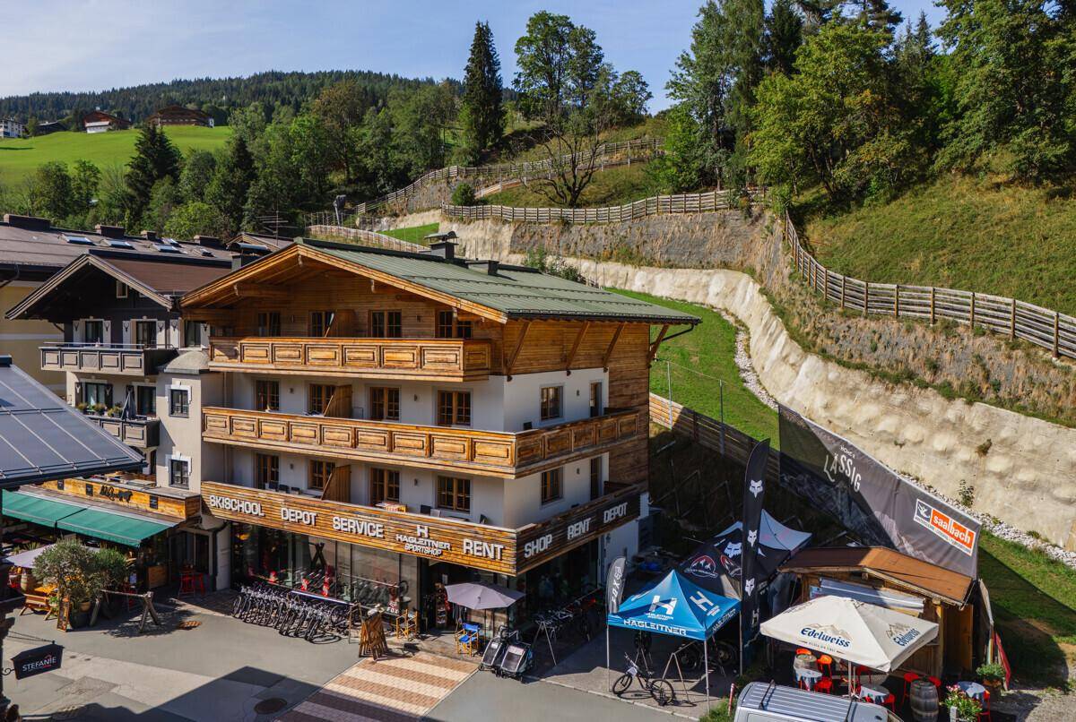 Alpines Gebäude mit Holzbalkonen und einem Sportgeschäft im Erdgeschoss, umgeben von grünen Hügeln.
