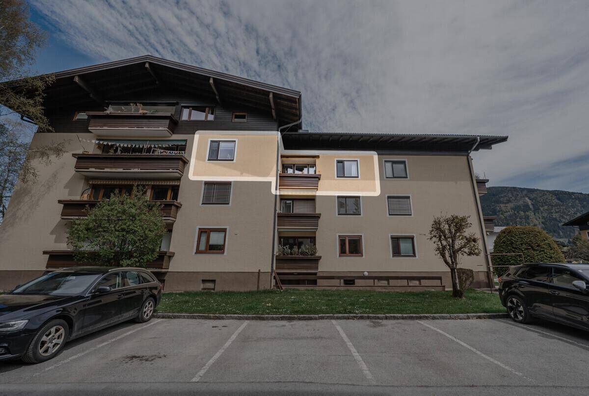 Mehrfamilienhaus mit Parkplätzen und Balkonen in alpiner Umgebung.