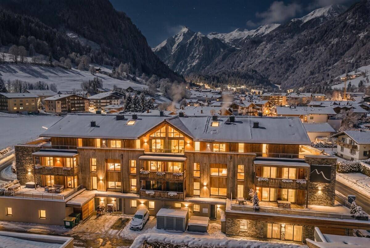 Beleuchtete Wohnanlage im alpinen Stil in einer verschneiten Berglandschaft bei Nacht.