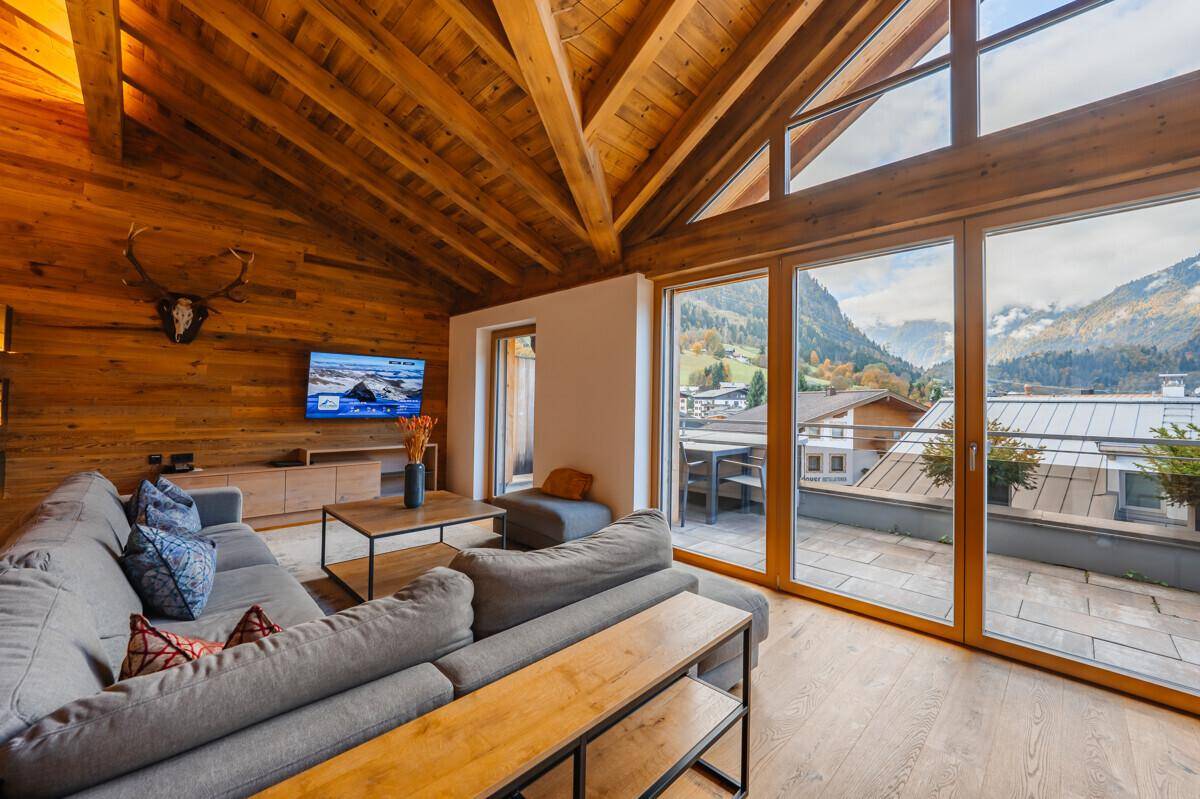 Helles Wohnzimmer mit Holzwänden, großem Sofa und Zugang zu einer Außenterrasse mit Bergblick.