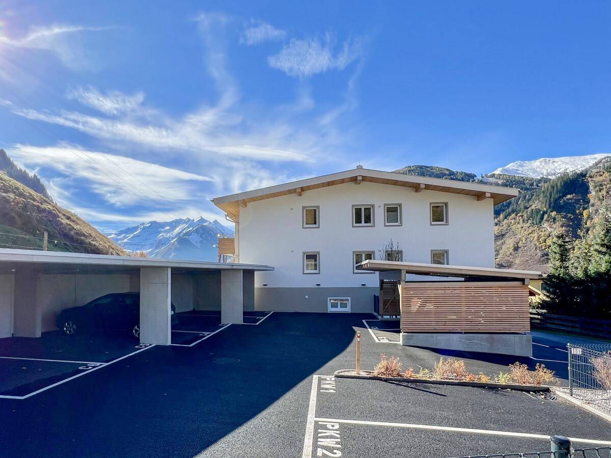 Modernes Gebäude in alpiner Landschaft mit Carport und Außenstellplätzen vor einer beeindruckenden Bergkulisse.