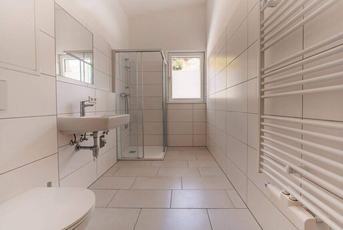 Modernes Badezimmer mit weißen Fliesen, Duschkabine, Waschbecken, WC und beheiztem Handtuchhalter.