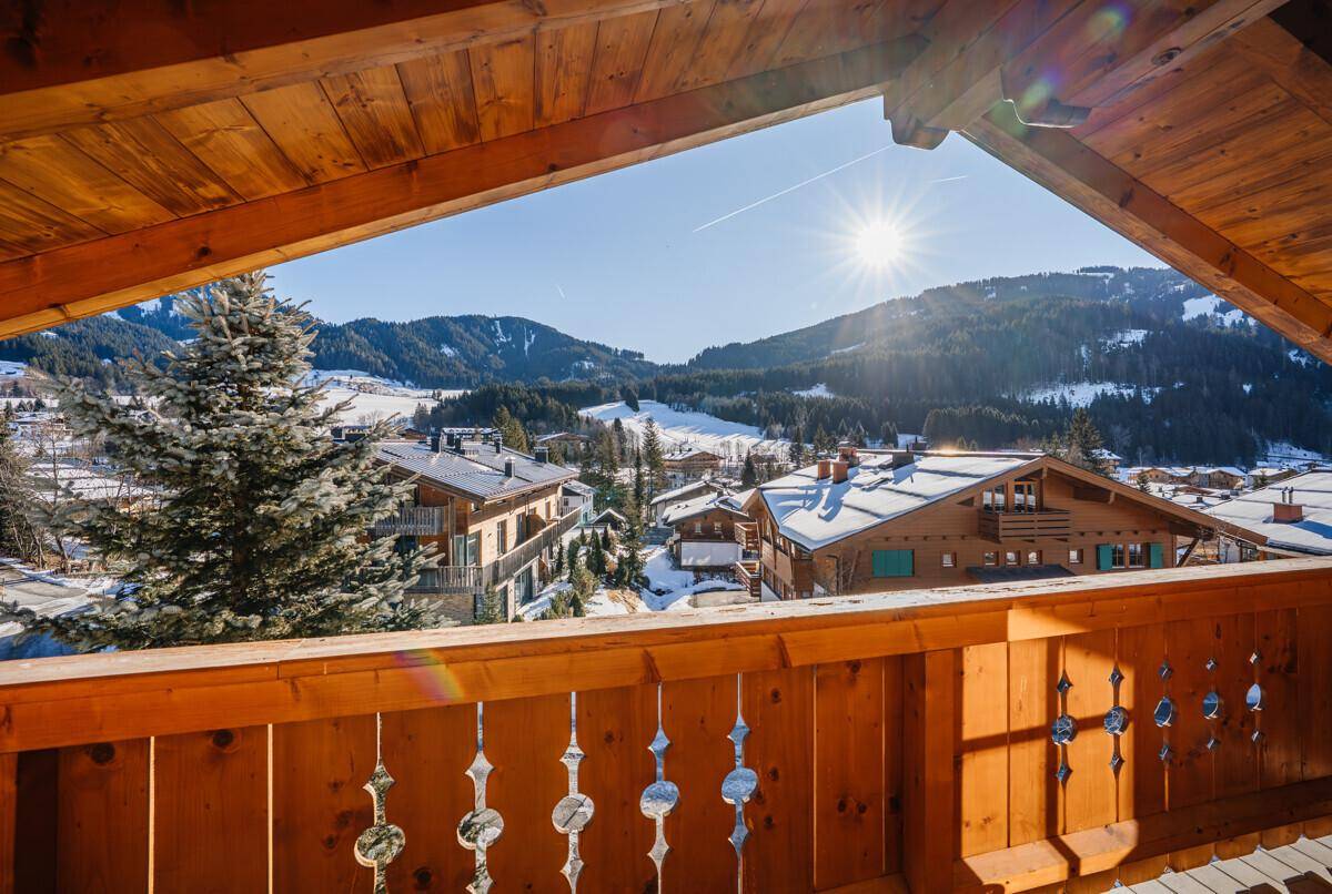 Sonniger Holzbalkon mit weitem Blick über die verschneite Alpenlandschaft und Dächer.
