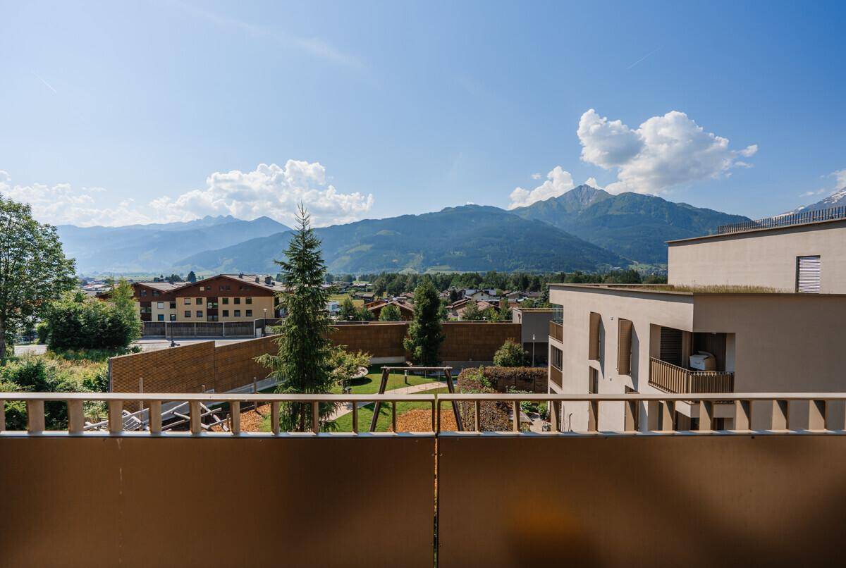 Weitläufiger Blick vom Balkon auf einen Spielplatz, grüne Bäume und die umliegende Berglandschaft.