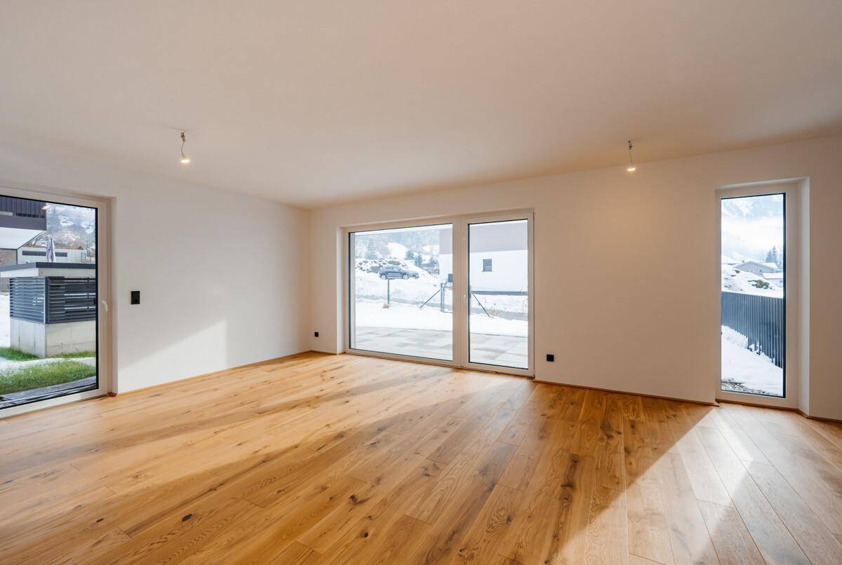 Helles, leeres Wohnzimmer mit großen Fenstern, Holzdielenboden und Blick auf die verschneite Landschaft.