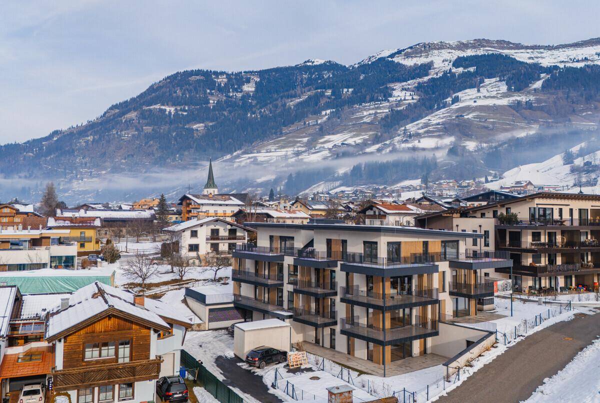 Apartmentgebäude im Winter mit verschneiter Umgebung und Blick auf ein Bergdorf.