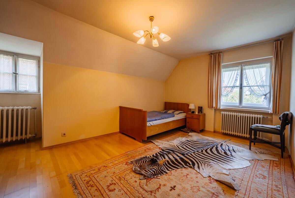 Geräumiges Schlafzimmer mit gelben Wänden, Parkettboden, zwei Fenstern und einem auffälligen Teppich.