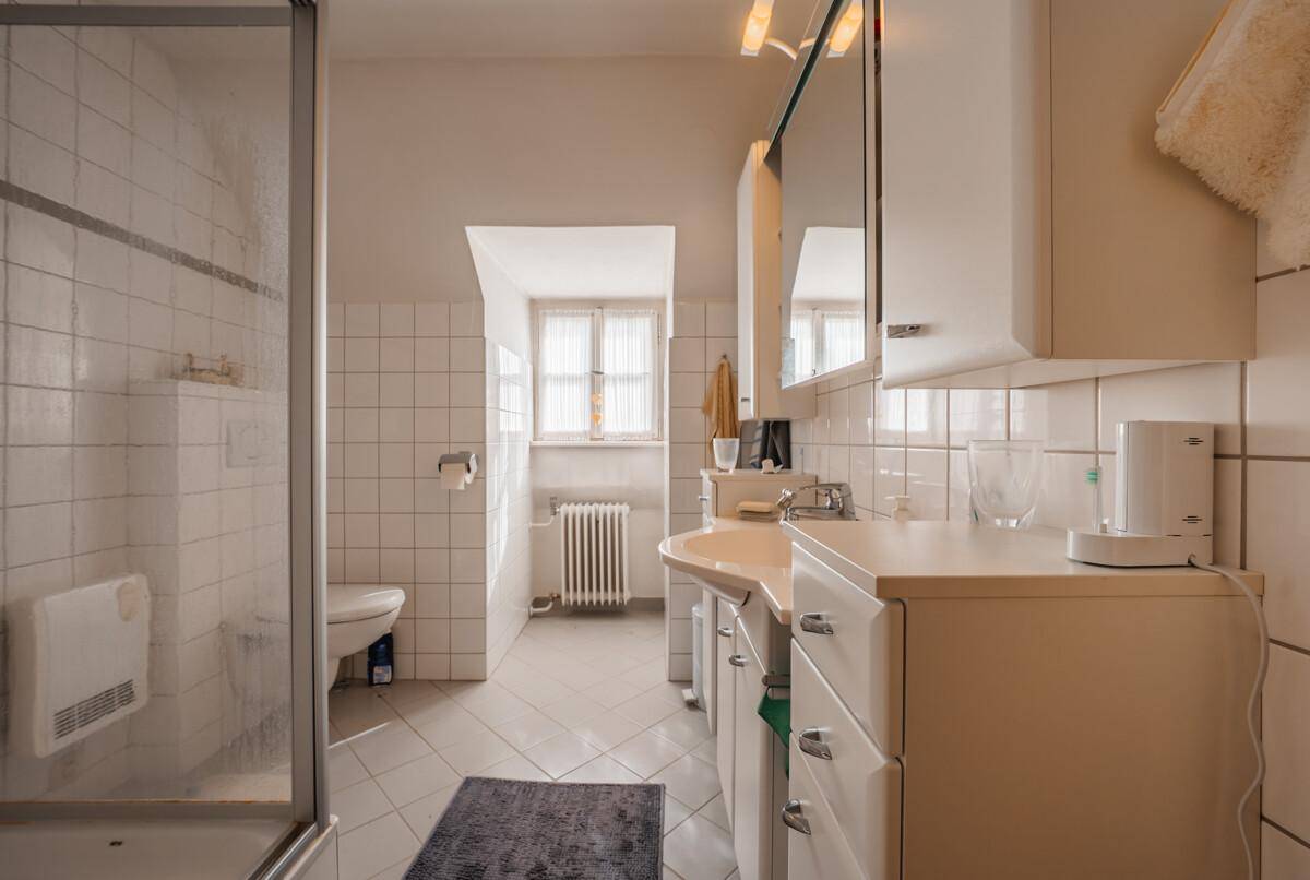 Helles Badezimmer mit weißen Fliesen, Dusche und einem Fenster für Tageslicht.