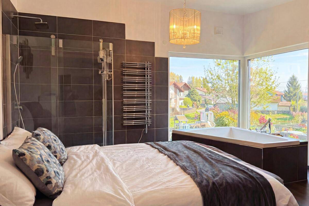Schlafzimmer mit integrierter Badewanne, Dusche und großen Panoramafenstern mit Blick ins Grüne.