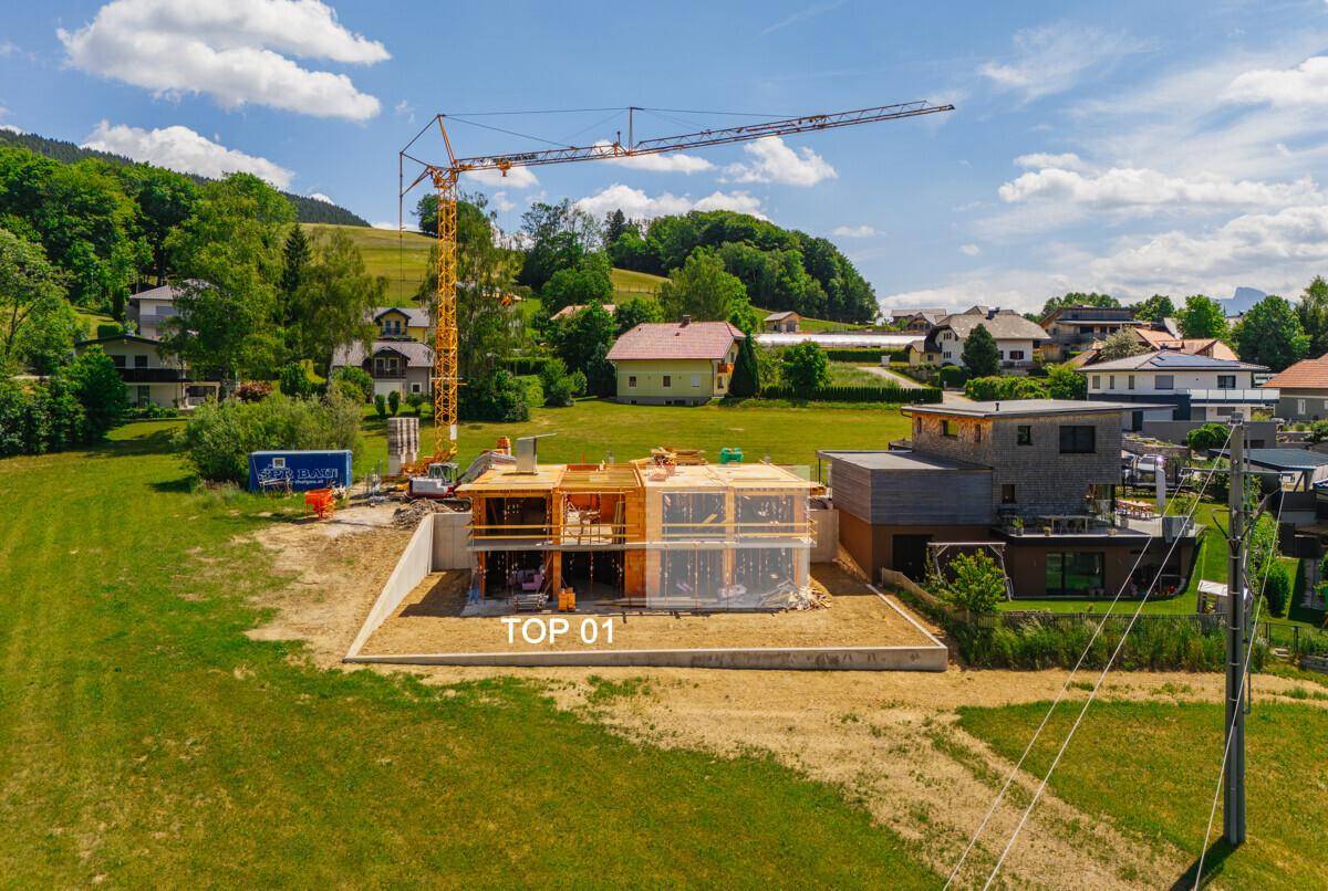Baustelle eines neuen Gebäudes mit Kran und teilweiser Struktur, umgeben von grüner Landschaft und Häusern.