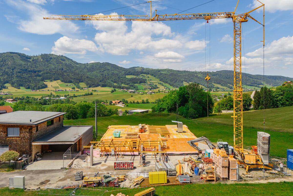 Baustelle mit Baumaterialien und einem großen Kran, umgeben von grünen Hügeln und ländlicher Idylle.