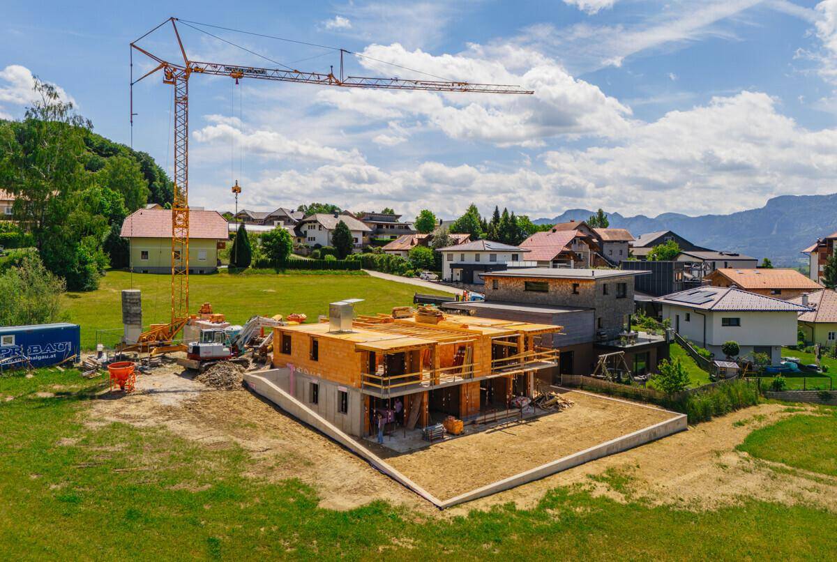 Baustelle eines Neubaus mit sichtbarem Kran und fortgeschrittener Holzrahmenkonstruktion in ländlicher Umgebung.