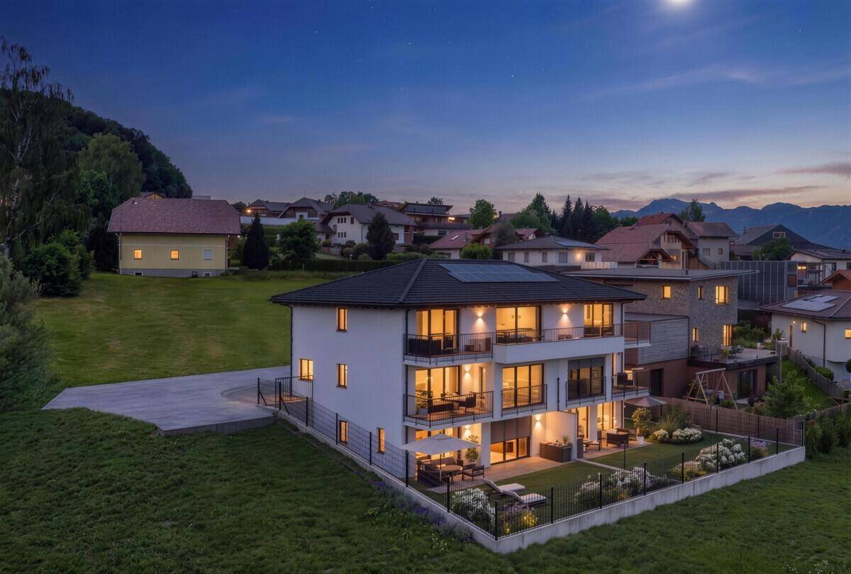Modernes Mehrfamilienhaus bei Dämmerung mit beleuchteten Innenräumen und Blick auf die umliegende Hügellandschaft.