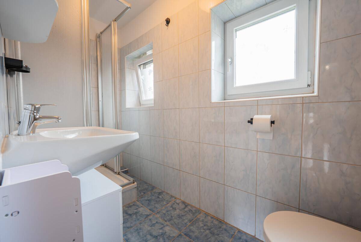 Funktionales Badezimmer mit hellblauen Fliesen, Dusche, WC und zwei Fenstern für Tageslicht.