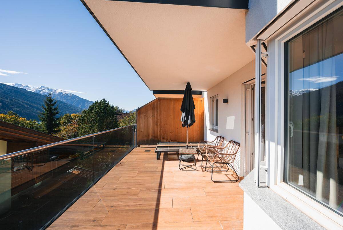 Großer Balkon mit Holzboden, Glasgeländer, Sitzgelegenheiten und Bergblick.