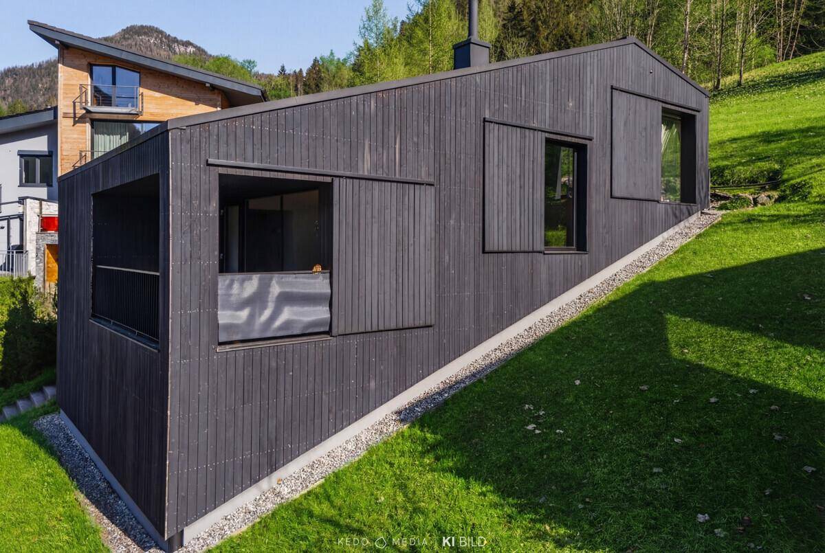 Moderne schwarze Holzfassade des Hauses mit großen Fenstern und angrenzendem Balkonbereich, umgeben von Grün.