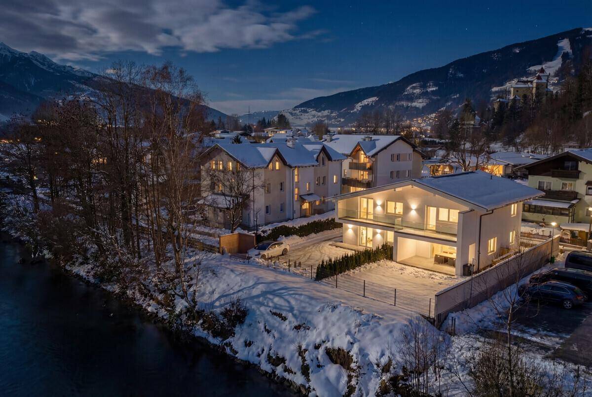 Abendliche Ansicht des beleuchteten Hauses im Winter mit verschneiter Umgebung und Bergen.