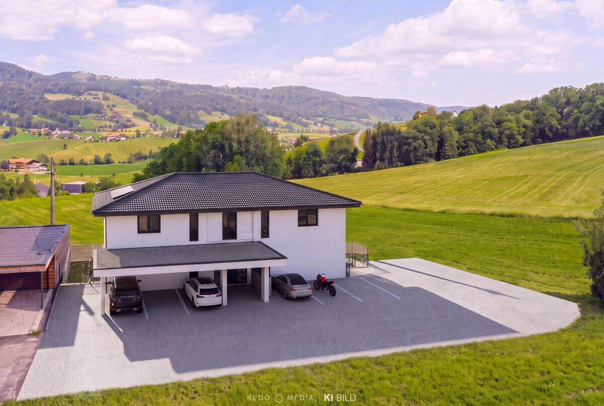 Modernes Wohngebäude mit Carport und Parkplätzen, umgeben von grünen Wiesen.