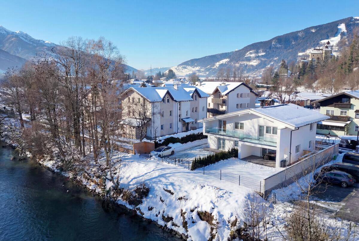 Winterliche Ansicht des modernen Hauses am Flussufer mit verschneiter Landschaft und Bergen.