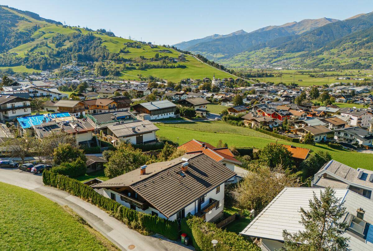 Luftaufnahme eines charmanten Dorfes eingebettet in grüne Hügel und majestätische Berge unter blauem Himmel.