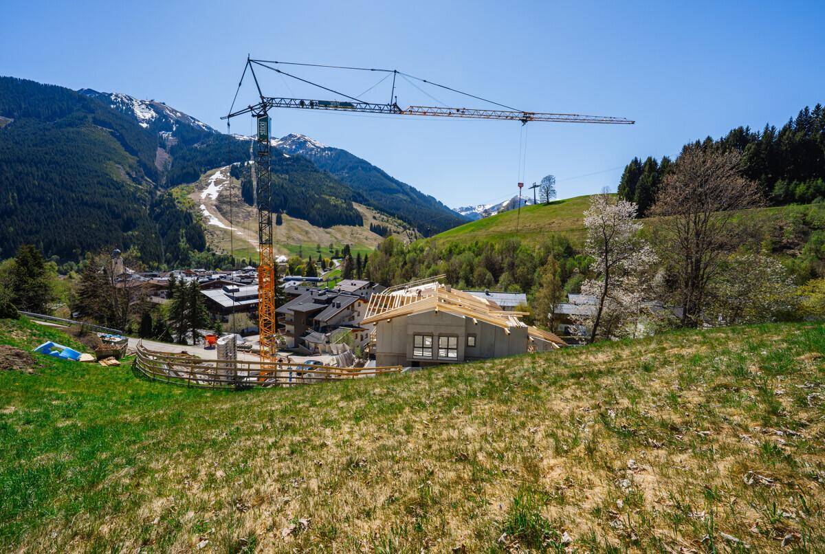 Baustelle mit Kran und einem im Bau befindlichen Haus vor einer beeindruckenden Bergkulisse.
